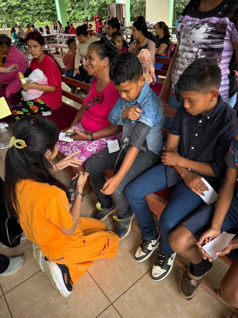 Children seated on benches receive medical attention from a healthcare worker wearing orange scrubs in a community setting.
