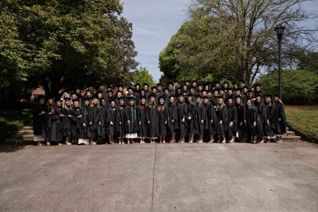 Large group of graduates in black gowns pose together outdoors in front of trees on a sunny day.