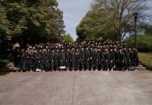 Physician assistant studies students earn perfect pass rate on national certifying exam Large group of graduates in black gowns pose together outdoors in front of trees on a sunny day.