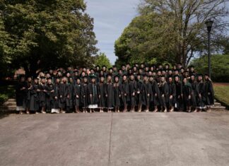 Physician assistant studies students earn perfect pass rate on national certifying exam Large group of graduates in black gowns pose together outdoors in front of trees on a sunny day.