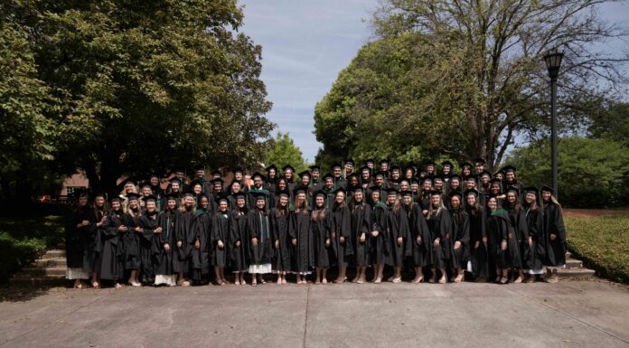 Physician assistant studies students earn perfect pass rate on national certifying exam Large group of graduates in black gowns pose together outdoors in front of trees on a sunny day.
