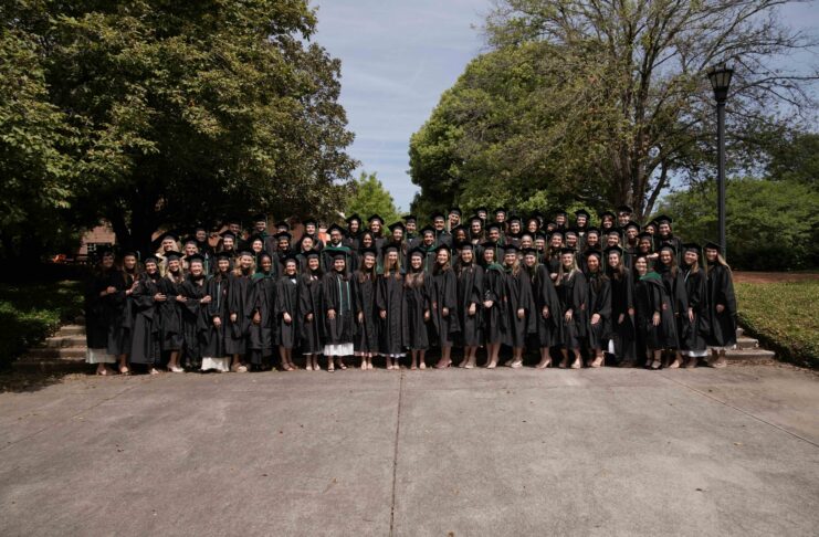 Large group of graduates in black gowns pose together outdoors in front of trees on a sunny day.