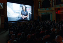 ‘Georgia Rising’ draws near-capacity crowd at Macon Film Festival, will air on GPB Audience watches a film in a theater showing a person in medical scrubs seated in a clinic, speaking to the camera.