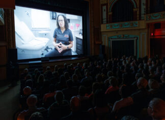 ‘Georgia Rising’ draws near-capacity crowd at Macon Film Festival, will air on GPB Audience watches a film in a theater showing a person in medical scrubs seated in a clinic, speaking to the camera.