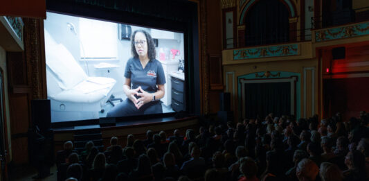 Audience watches a film in a theater showing a person in medical scrubs seated in a clinic, speaking to the camera.