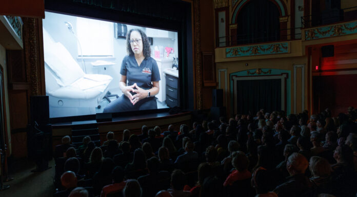 ‘Georgia Rising’ draws near-capacity crowd at Macon Film Festival, will air on GPB Audience watches a film in a theater showing a person in medical scrubs seated in a clinic, speaking to the camera.