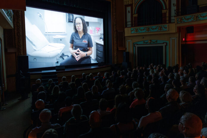 2026GeorgiaRisingJohnKnight00201 Audience watches a film in a theater showing a person in medical scrubs seated in a clinic, speaking to the camera.
