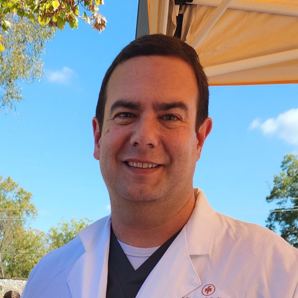 Man wearing a white coat and standing outside under a canopy with trees and blue sky in the background.
