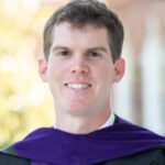 Young man wearing a graduation gown and purple hood, smiling outdoors with blurred background.