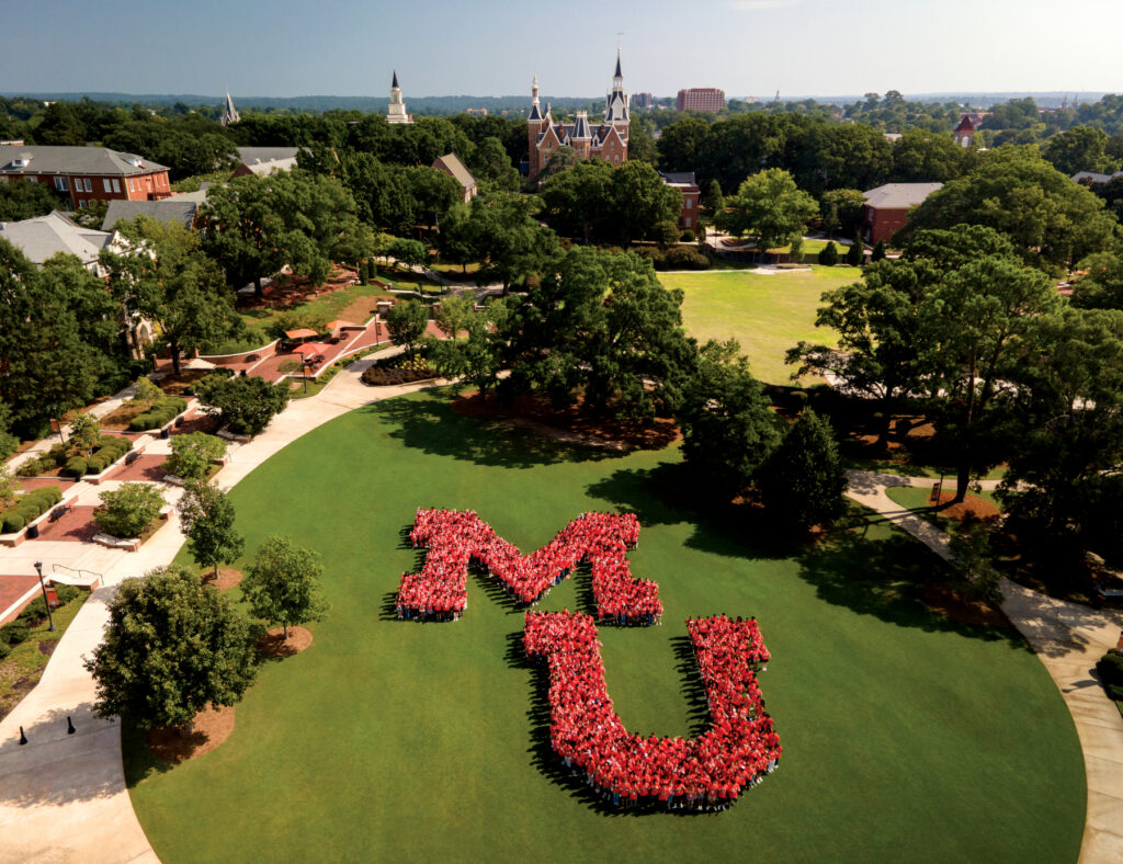 Students form a large MU on a campus lawn, with historic university buildings and trees in the background on a sunny day.