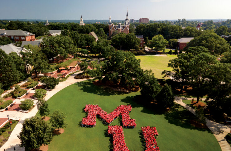 Students form a large MU on Cruz Plaza, with historic university buildings and trees in the background on a sunny day.
