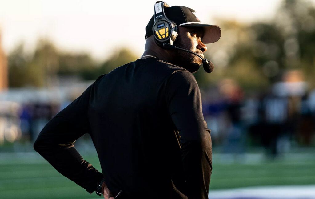 Football coach wearing a headset stands on the field with hands on hips, looking toward the game.