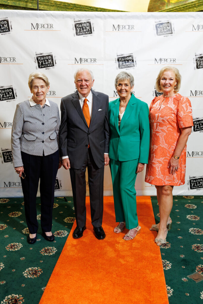 Four adults pose on an orange carpet in front of a step-and-repeat banner at an event.