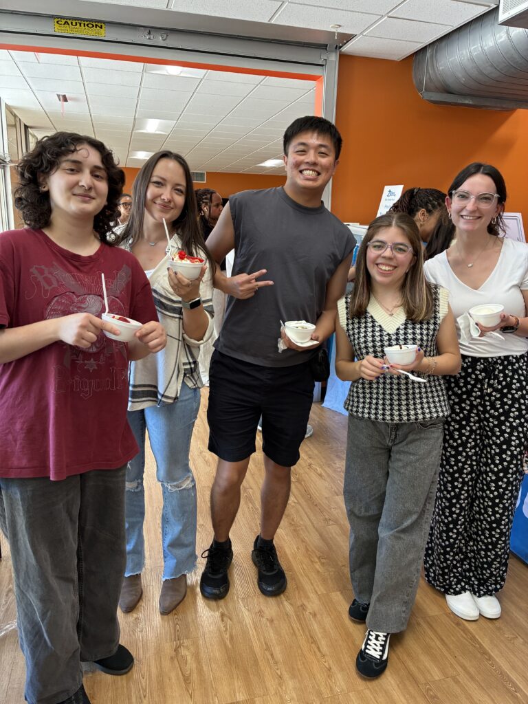 Five people stand indoors, smiling and holding bowls of ice cream, with others and bright walls in the background.
