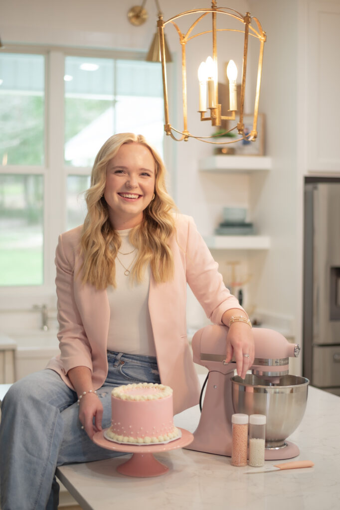 Woman smiles in a kitchen, sitting beside a pink cake and matching stand mixer on the counter.