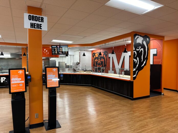 Fast food restaurant interior with self-order kiosks, serving counter, and bear mascot wall art.