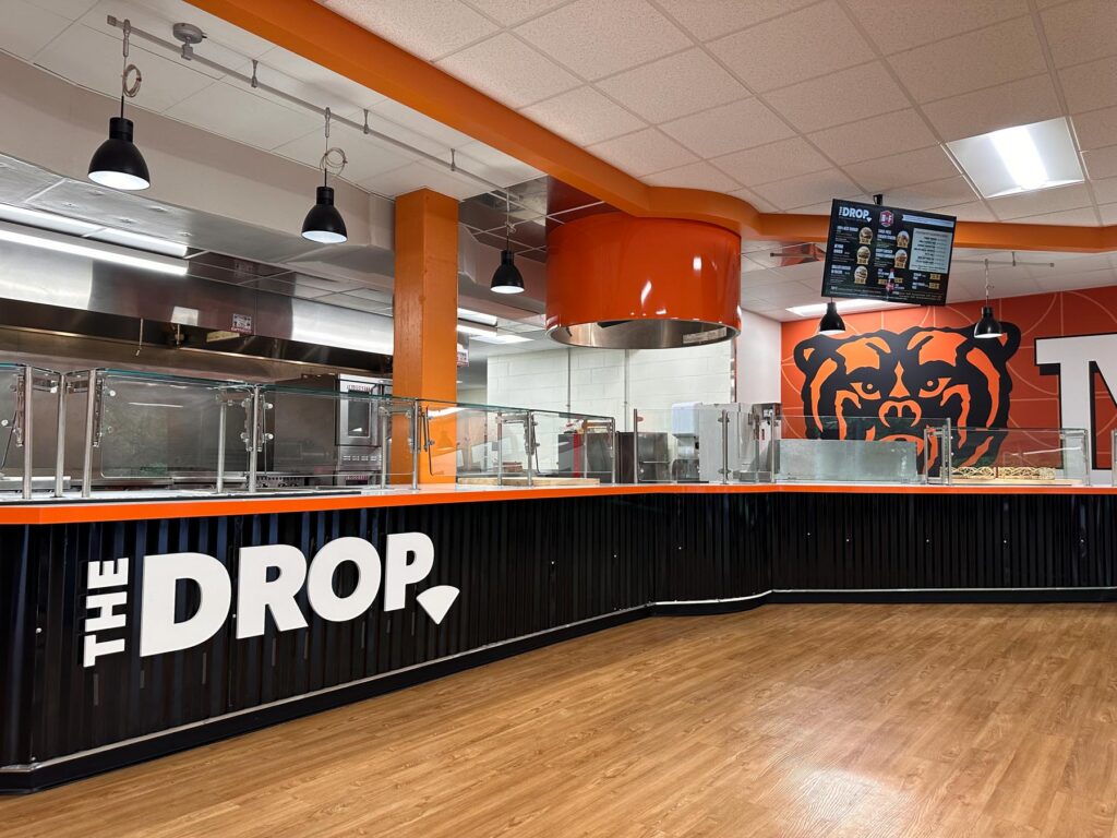 Empty cafeteria counter labeled The Drop, with a bear mural and menu screens in the background.
