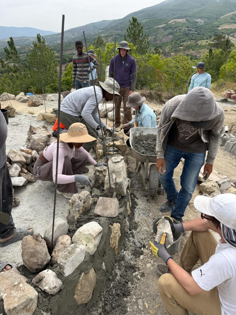 A group of people build a stone and concrete wall outdoors, surrounded by trees and hills in the background.