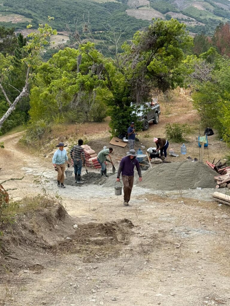 A group of people work outdoors mixing cement and moving bags near trees and a parked vehicle on a dirt road.