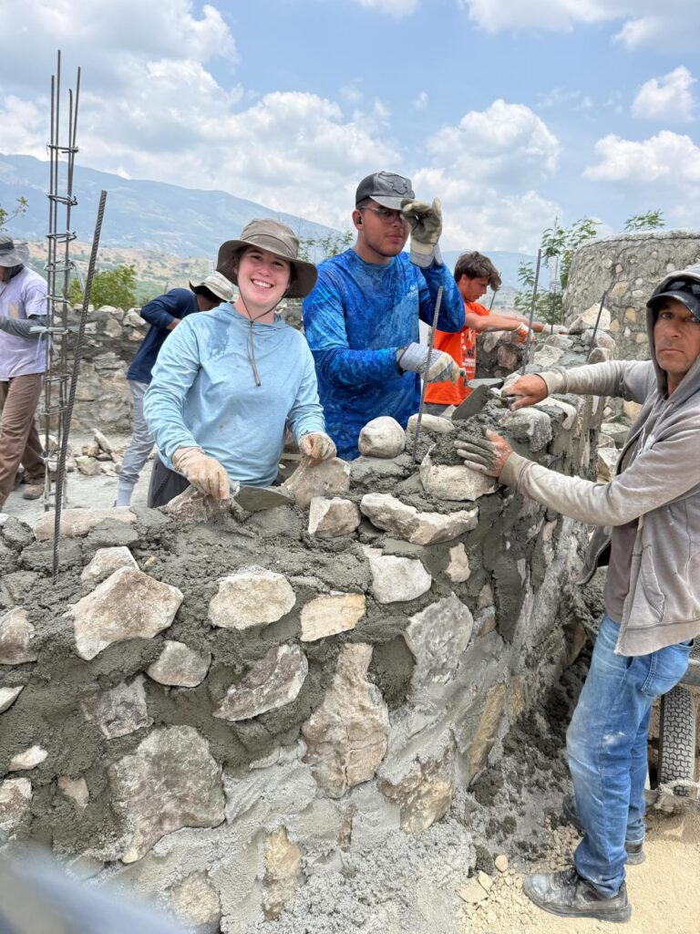 Several people work together building a stone wall outdoors, handling rocks and cement under a partly cloudy sky.