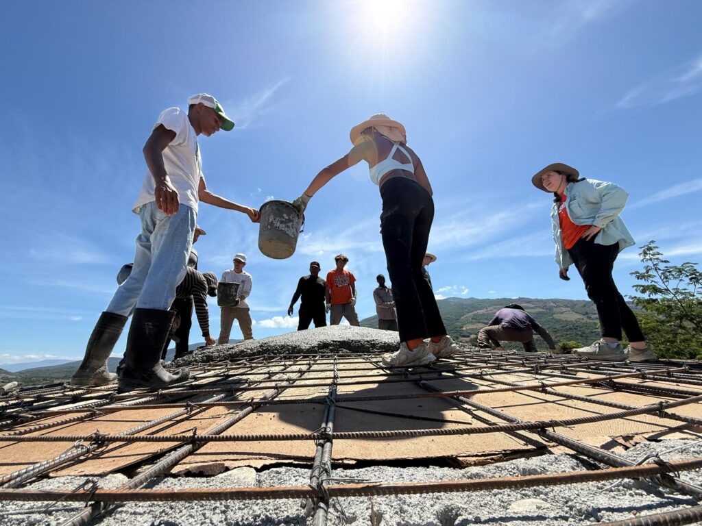 People work together pouring concrete on a rebar structure under a bright sun with clear blue skies in the background.