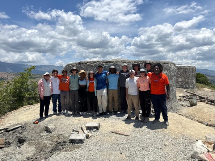 A group of people stands in front of a stone water tank under a partly cloudy sky in a rural, hilly landscape.