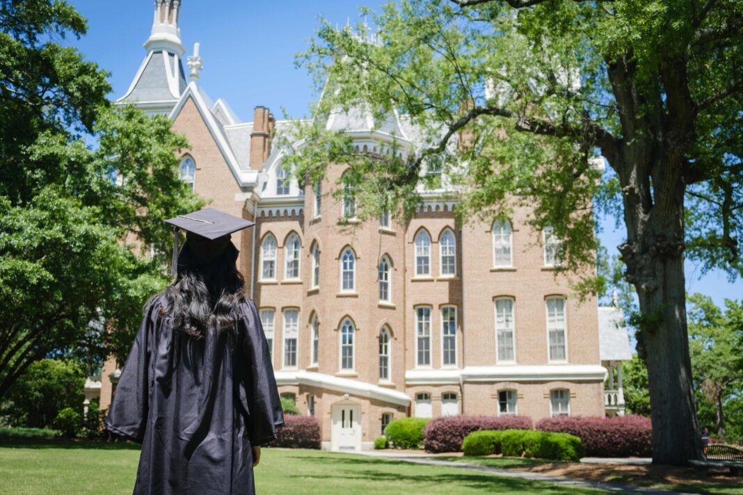 A graduate in cap and gown stands on grass facing a large, historic brick building surrounded by trees.