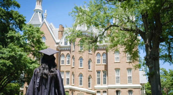 Mercer one of Princeton Review’s ‘Best Colleges’ for 24th consecutive year A graduate in cap and gown stands on grass facing a large, historic brick building surrounded by trees.