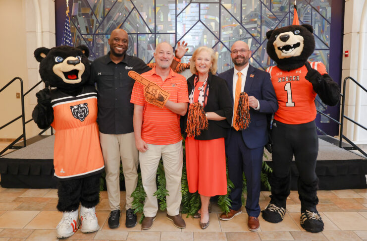 Five people, including two bear mascots, pose on stage with pom-poms and a foam finger at an indoor event.
