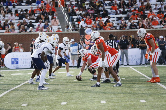 Two college football teams line up at the line of scrimmage during a daytime game, with fans watching in the stands.