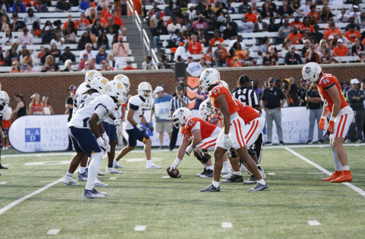 Two college football teams line up at the line of scrimmage during a daytime game, with fans watching in the stands.