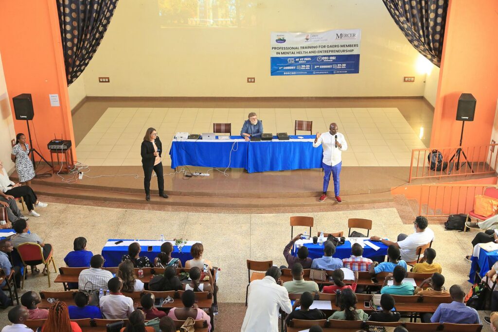 A speaker addresses an audience in a seminar room, with two people seated at a table on stage and a banner in the background.