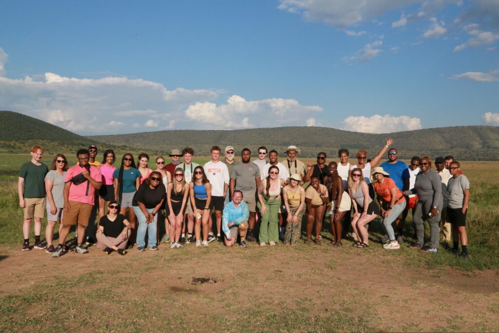 A group of people pose for a photo outdoors on grass with hills and a partly cloudy sky in the background.