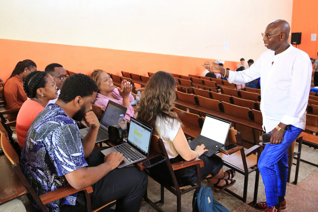 A group of students with laptops listen to an instructor speaking in a lecture hall with orange walls.