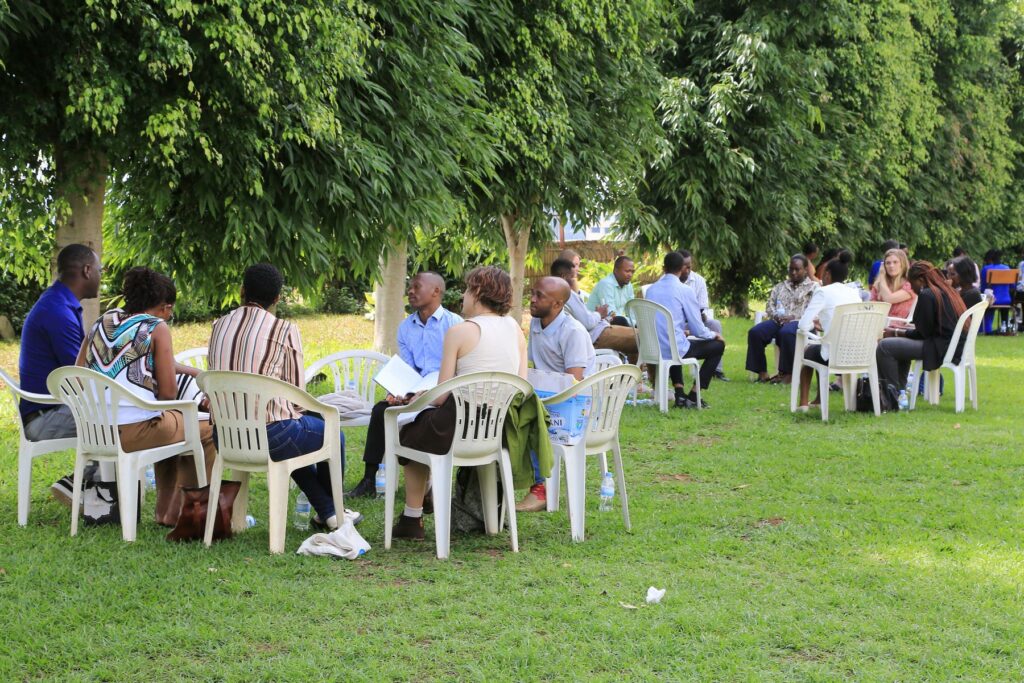 People sitting in small groups on plastic chairs outdoors, participating in a discussion or meeting under trees.