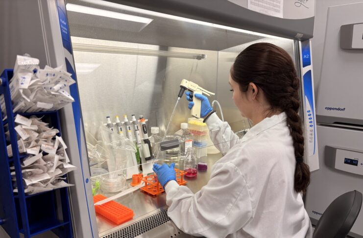 Scientist in lab coat and gloves uses pipette at biosafety cabinet with various lab supplies and containers.