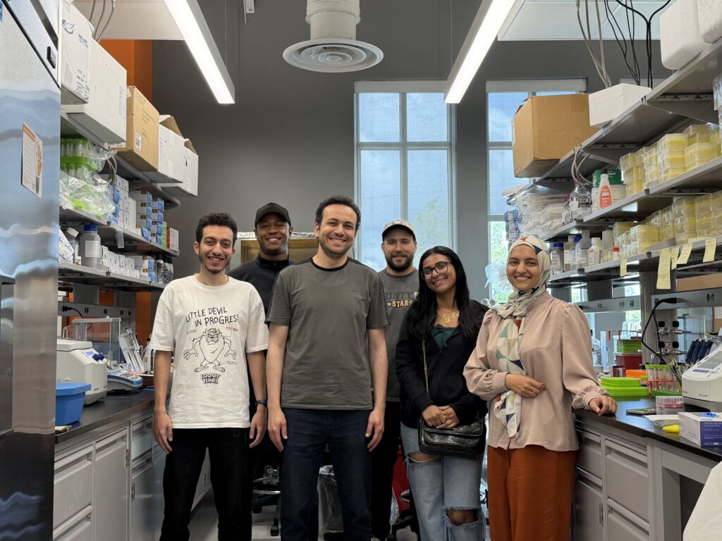 Six people standing and smiling inside a modern laboratory with shelves of equipment and supplies visible around them.