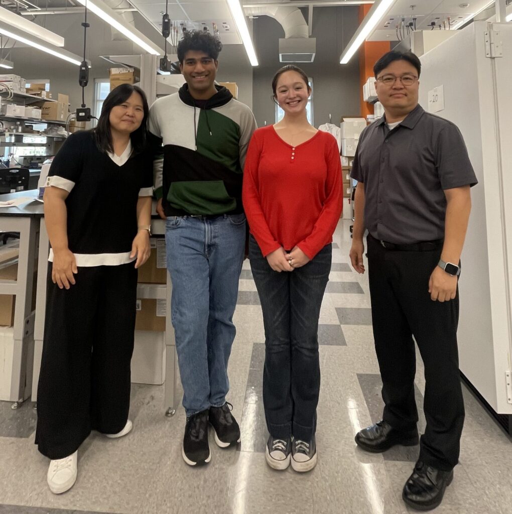 Four people standing and smiling in a lab setting with equipment and shelves visible in the background.