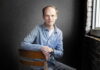English associate professor awarded prestigious Amy Lowell Traveling Poetry Scholarship A man in a light blue shirt sits on a chair in front of a dark background near a window with brick walls.