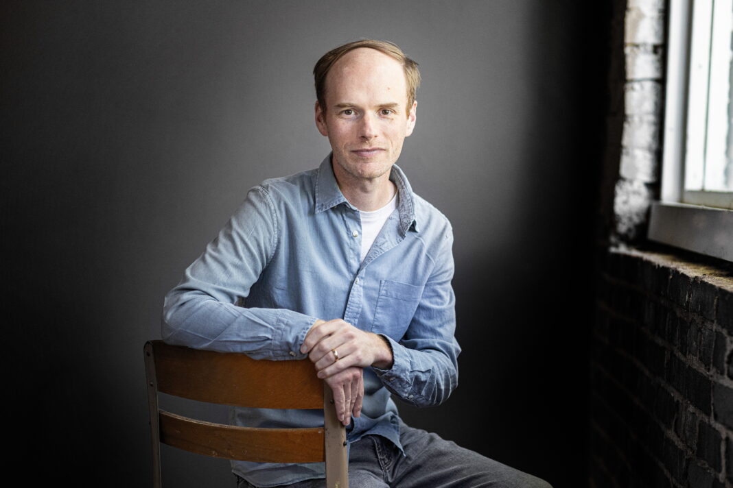 A man in a light blue shirt sits on a chair in front of a dark background near a window with brick walls.