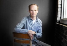 English associate professor awarded prestigious Amy Lowell Traveling Poetry Scholarship A man in a light blue shirt sits on a chair in front of a dark background near a window with brick walls.