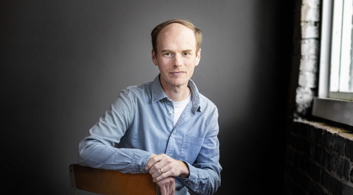 English associate professor awarded prestigious Amy Lowell Traveling Poetry Scholarship A man in a light blue shirt sits on a chair in front of a dark background near a window with brick walls.