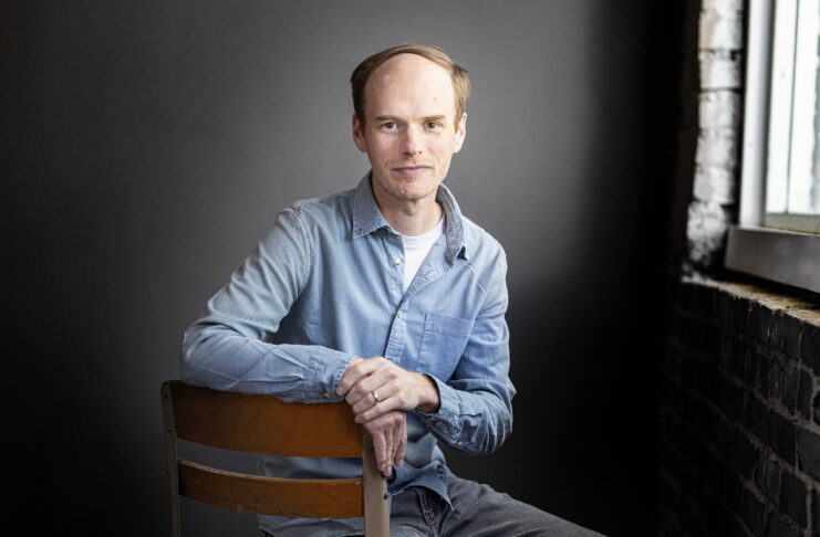A man in a light blue shirt sits on a chair in front of a dark background near a window with brick walls.
