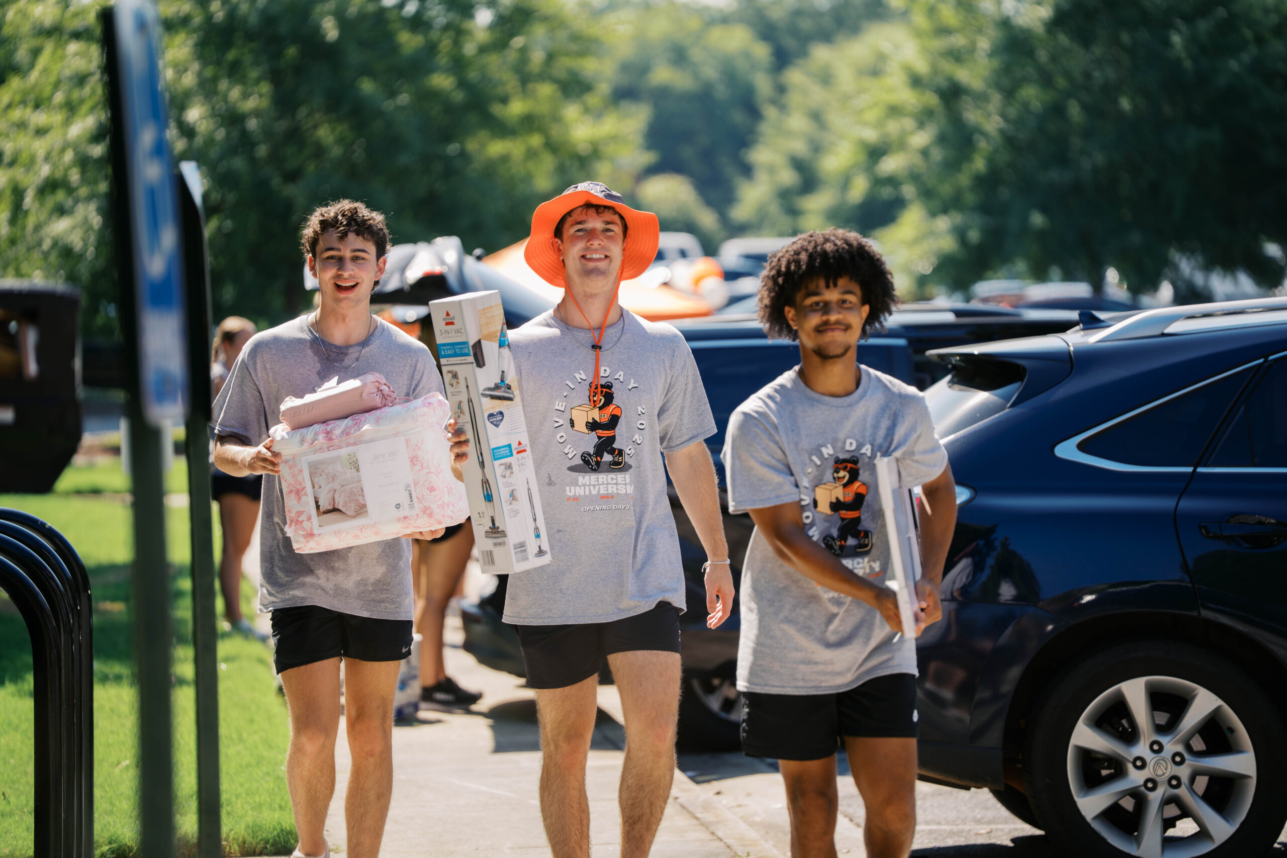Mercer to Welcome Class of 2030 for Opening Days Three people in matching t-shirts and shorts carry supplies on a sunny day near parked cars and greenery.
