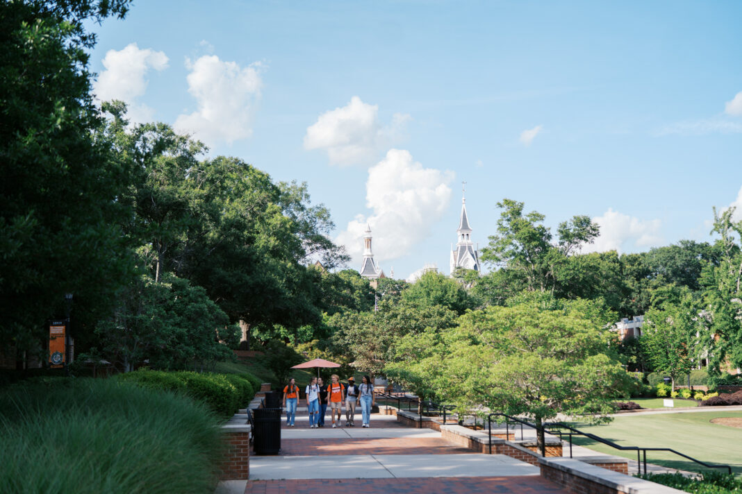 Four people walk on a tree-lined brick path with two white church steeples visible in the background under a blue sky.