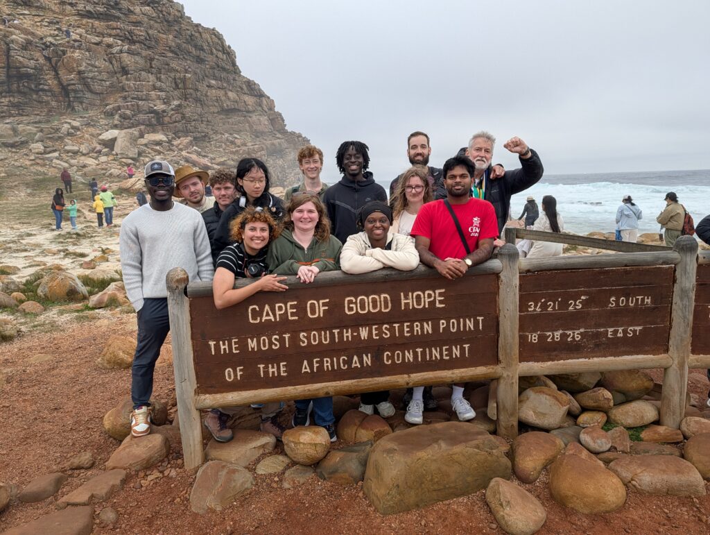 A group of people pose by a sign marking the Cape of Good Hope in South Africa with rocks and ocean in the background.
