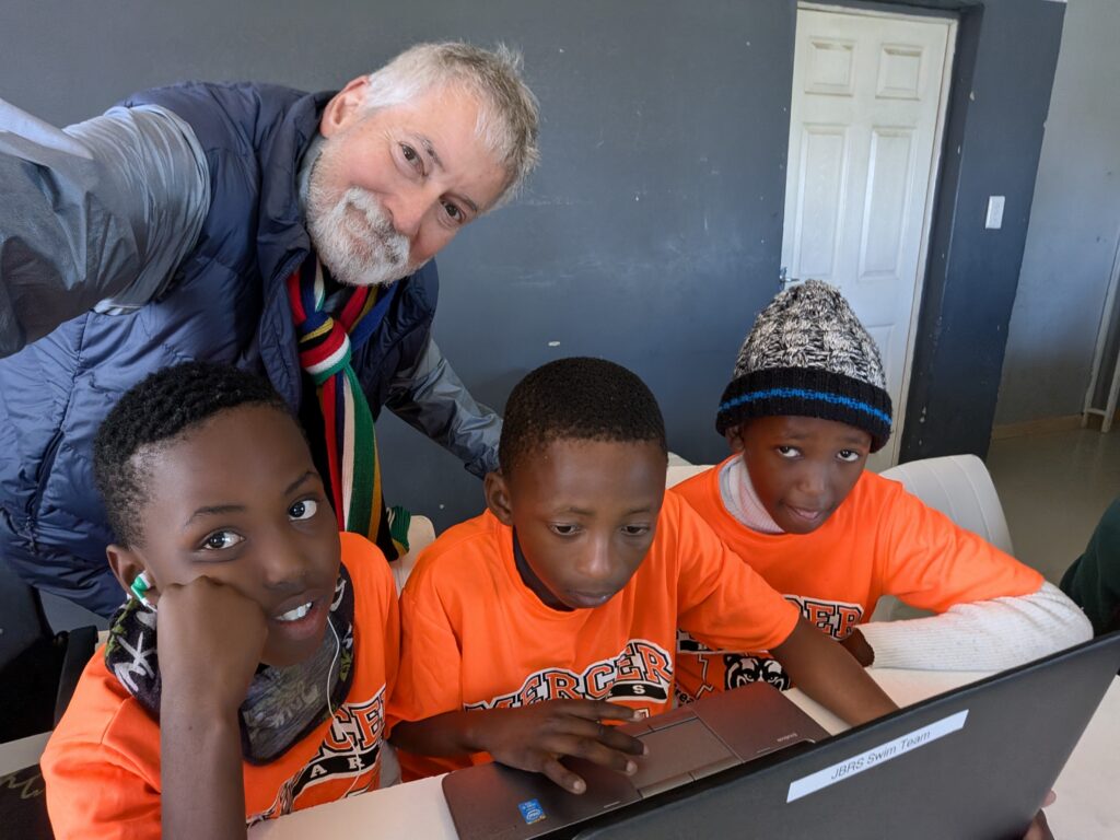 Three boys in orange shirts use a laptop at a table while an older man stands behind them, smiling at the camera.