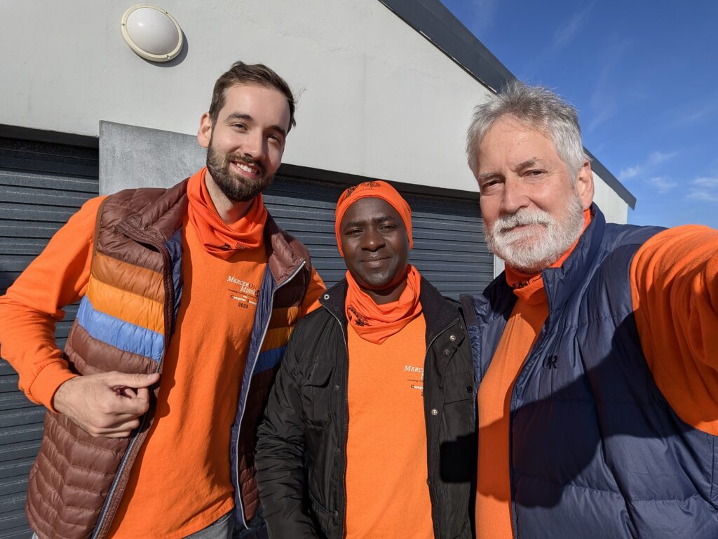 Three men wearing orange shirts and jackets stand outside in sunlight, posing for a group photo and smiling.
