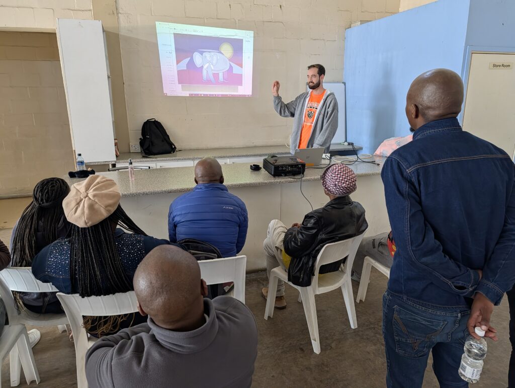 A man gives a presentation to a seated group, showing an elephant illustration on a projected screen.
