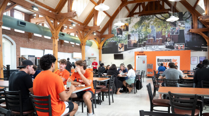 What’s new with Mercer dining for fall 2026 Students sit and talk in a busy university dining hall with wooden beams and a large mural on the back wall.